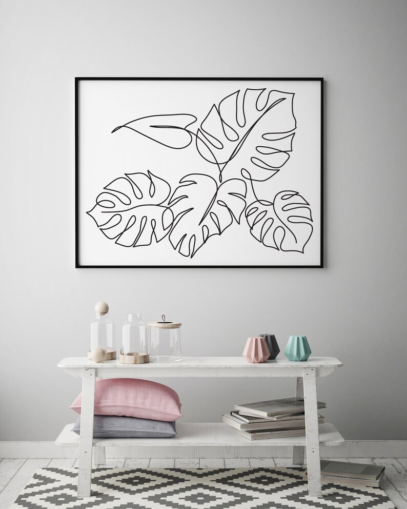 May include: A black and white line drawing of tropical leaves in a black frame hanging on a white wall above a white shelf with two clear glass jars, a wooden knob, and three colorful ceramic candle holders.
