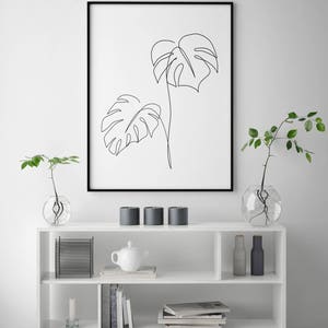 May include: A black framed print of a line drawing of two monstera leaves on a white background. The print is hanging on a white wall above a white shelf with a white teapot, candles, and books.