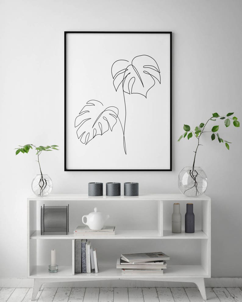 May include: A black framed print of a line drawing of two monstera leaves on a white background. The print is hanging on a white wall above a white shelf with a white teapot, candles, and books.