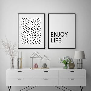 May include: Two framed art prints. One features a pattern of black dots on a white background. The other print has the words "ENJOY LIFE" in black text on a white background. The prints are displayed above a white dresser.