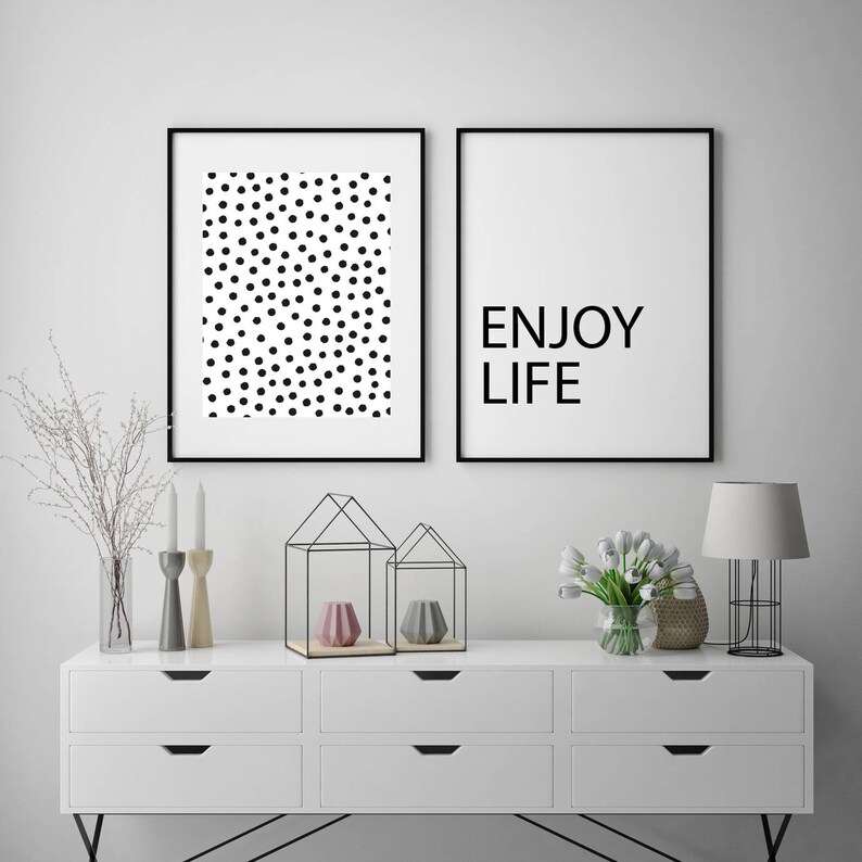 May include: Two framed art prints. One features a pattern of black dots on a white background. The other print has the words "ENJOY LIFE" in black text on a white background. The prints are displayed above a white dresser.
