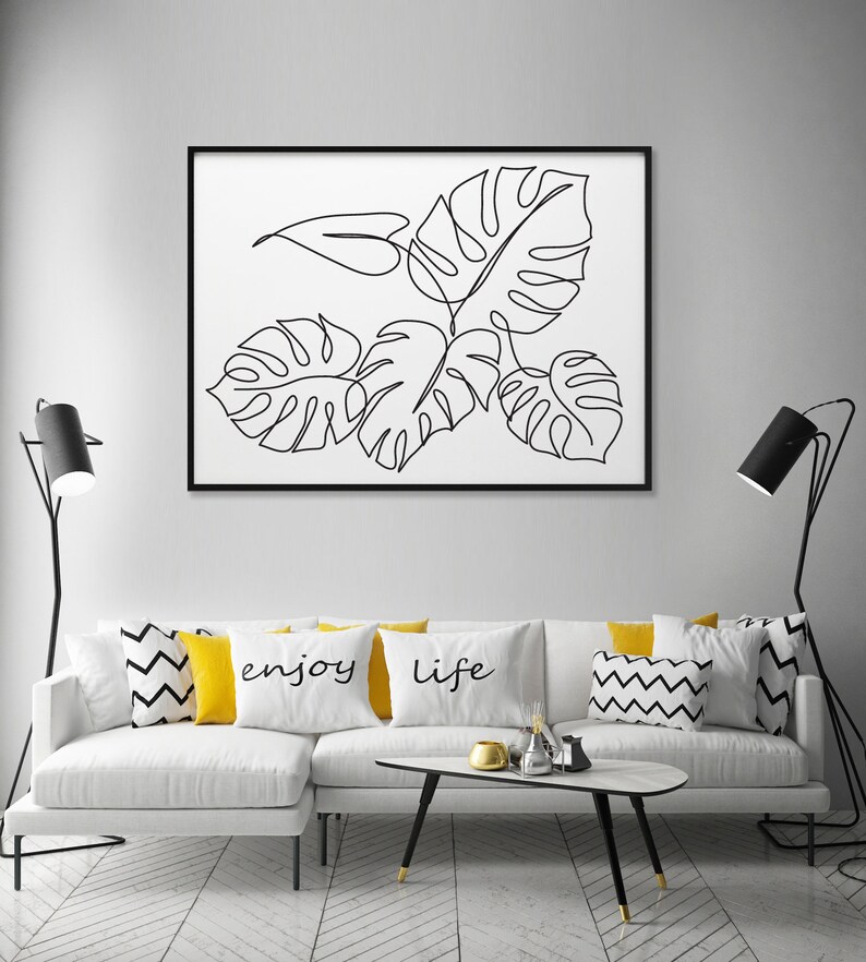 May include: A white couch with yellow and black patterned throw pillows. The pillows have the words "enjoy" and "life" printed on them. A black and white line drawing of tropical leaves is framed and hanging on the wall above the couch.