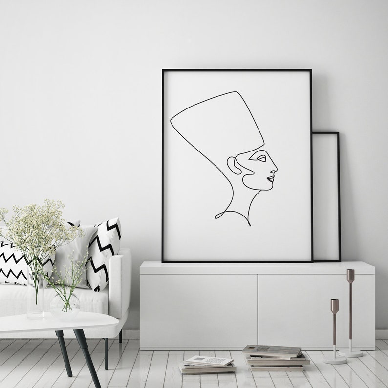 May include: A black and white line drawing of a woman wearing a headdress, in a minimalist style, framed in a black frame.
