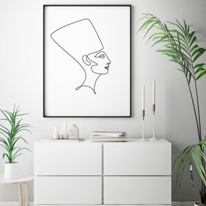 May include: A black and white line drawing of a woman wearing a headdress, framed in a black frame and hanging on a white wall above a white dresser with two drawers.