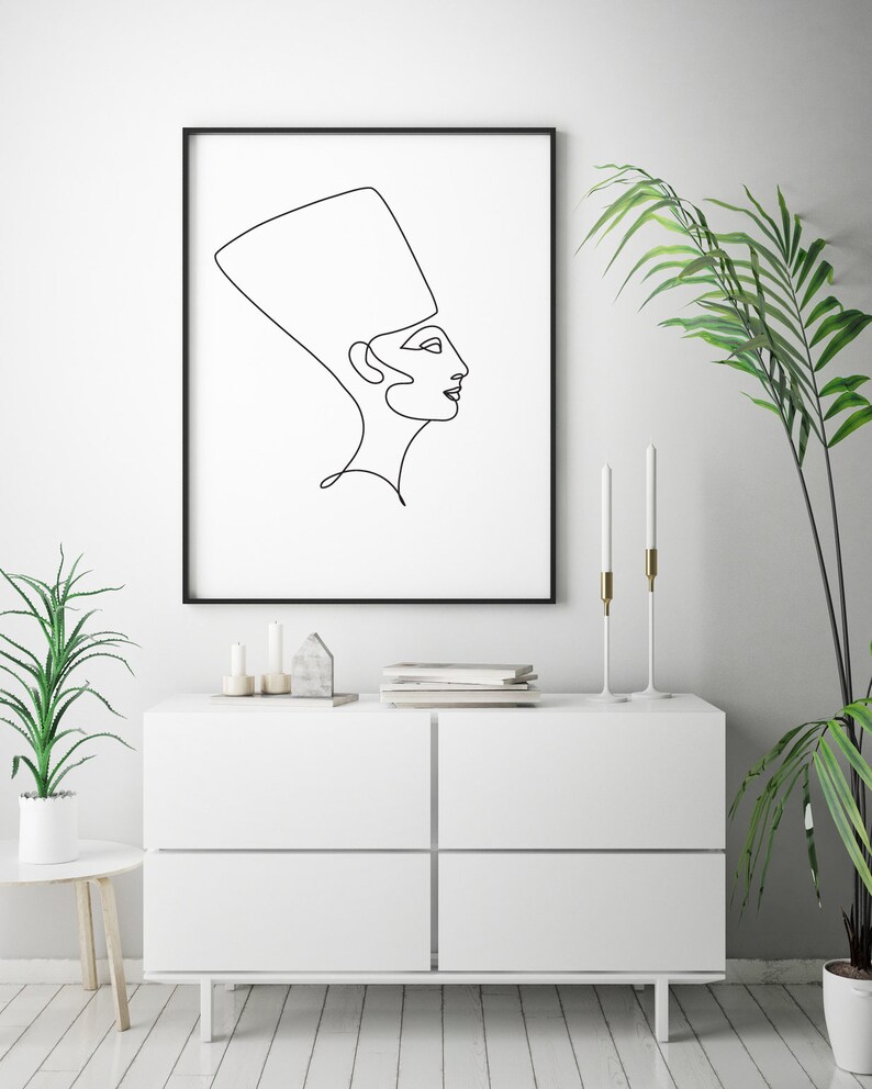 May include: A black and white line drawing of a woman wearing a headdress, framed in a black frame and hanging on a white wall above a white dresser with two drawers.