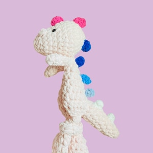 May include: A crochet dinosaur plush toy made of soft, light pink yarn. The creature features a row of colorful spikes along its back, transitioning from dark blue to light blue and mint green, with two bright pink spikes on its head.