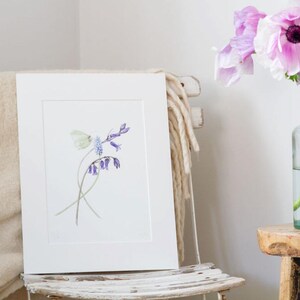 May include: A white matted print of a watercolor painting of bluebells and a white flower. The print is framed and displayed on a white chair.