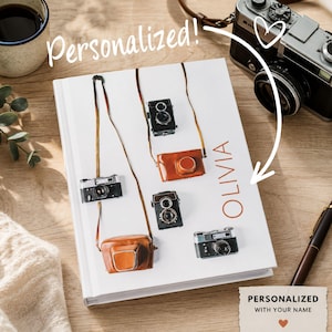 Op de afbeelding: Een wit notitieboek met harde kaft voorzien van een printpatroon van diverse vintage camera's met bruine lederen hoesjes en riemen. De naam OLIVIA staat in oranje verticale tekst gedrukt. Een wit briefje vermeldt PERSONALIZED WITH YOUR NAME.