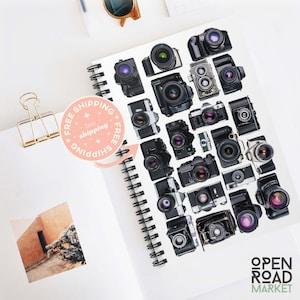 May include: A white notebook with a black spiral binding. The notebook is covered with black and silver vintage camera stickers. The text "OPEN ROAD MARKET" is printed in green on the bottom right corner of the notebook.