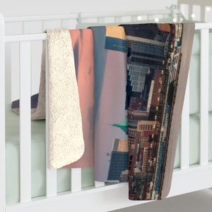 May include: A soft, plush throw blanket with a cityscape design. The blanket features a view of a city skyline at sunset with a pink and purple sky. The blanket is made of a soft, comfortable material and is perfect for cuddling up on the couch.