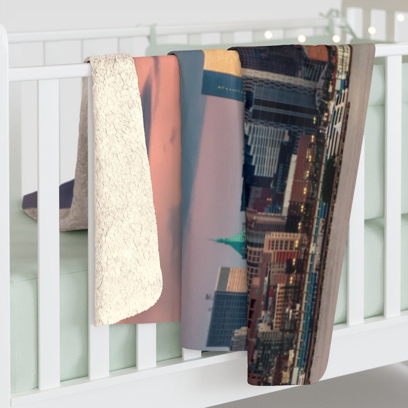 May include: A soft, plush throw blanket with a cityscape design. The blanket features a view of a city skyline at sunset with a pink and purple sky. The blanket is made of a soft, comfortable material and is perfect for cuddling up on the couch.