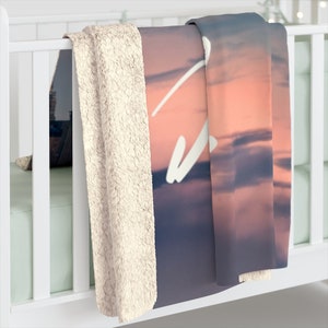 May include: A soft, plush fleece blanket with a pink and purple sunset sky design. The blanket has a white fluffy backing.