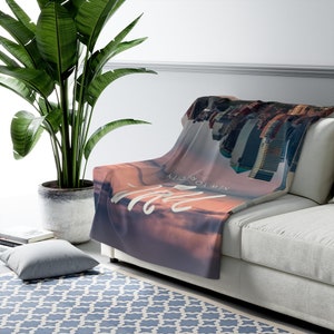 May include: A soft, plush throw blanket featuring a cityscape with a pink and purple sunset sky. The text "NEW TO CITY & BACK" is written in white on the blanket.