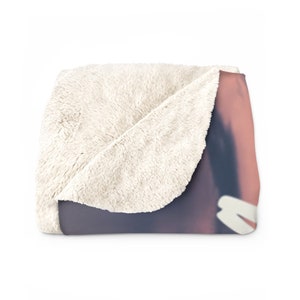 May include: A soft, white fleece blanket with a pink and purple gradient design on the front. The blanket is folded in half, showing the white fleece on the back.