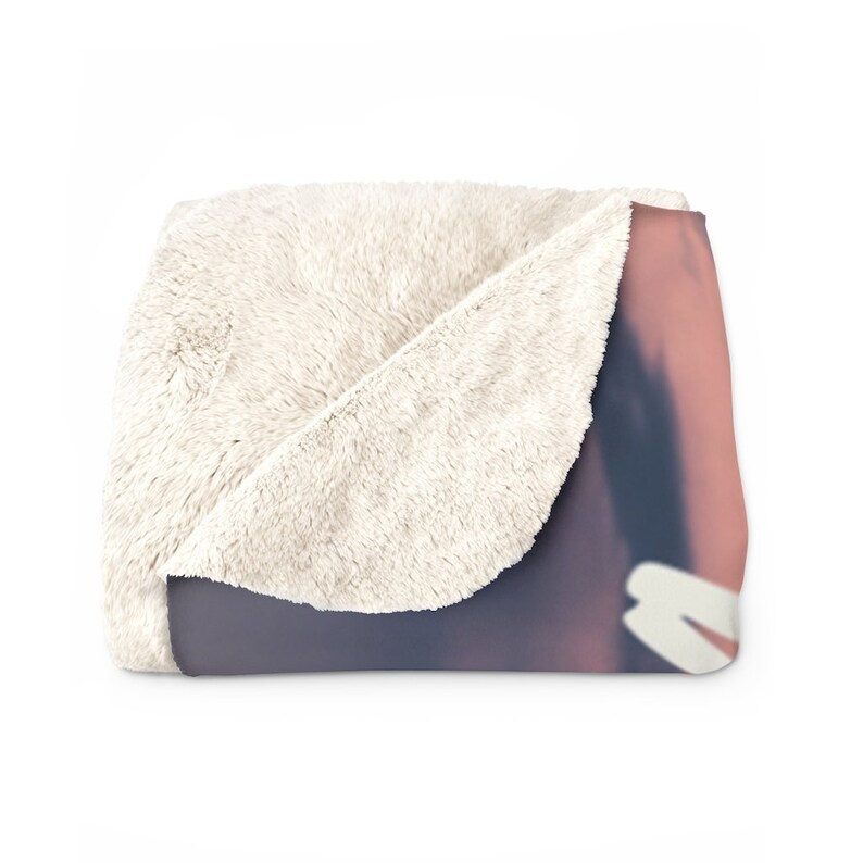 May include: A soft, white fleece blanket with a pink and purple gradient design on the front. The blanket is folded in half, showing the white fleece on the back.