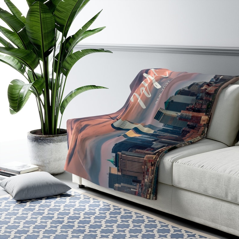 May include: A soft, plush throw blanket featuring a cityscape of New York City at sunset with the text "New York" in white script. The blanket is draped over a white couch.