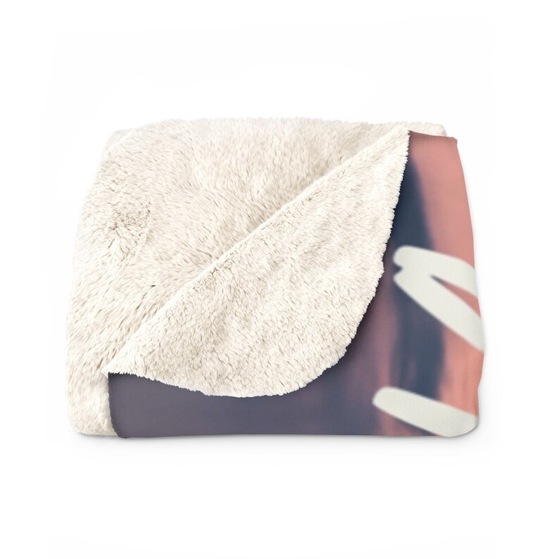 May include: A soft, white faux fur throw blanket with a pink and purple sunset printed on the front. The blanket is folded in half, showing the white faux fur on the back.