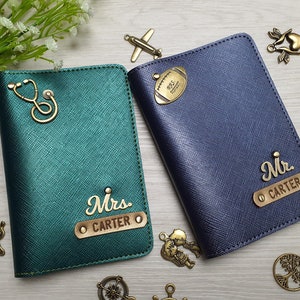 May include: Two personalized passport holders. One is teal green with a stethoscope charm and "Mrs. CARTER" in gold lettering. The other is navy blue with a football charm and "Mr. CARTER" in gold lettering. Decorative charms surround the holders.