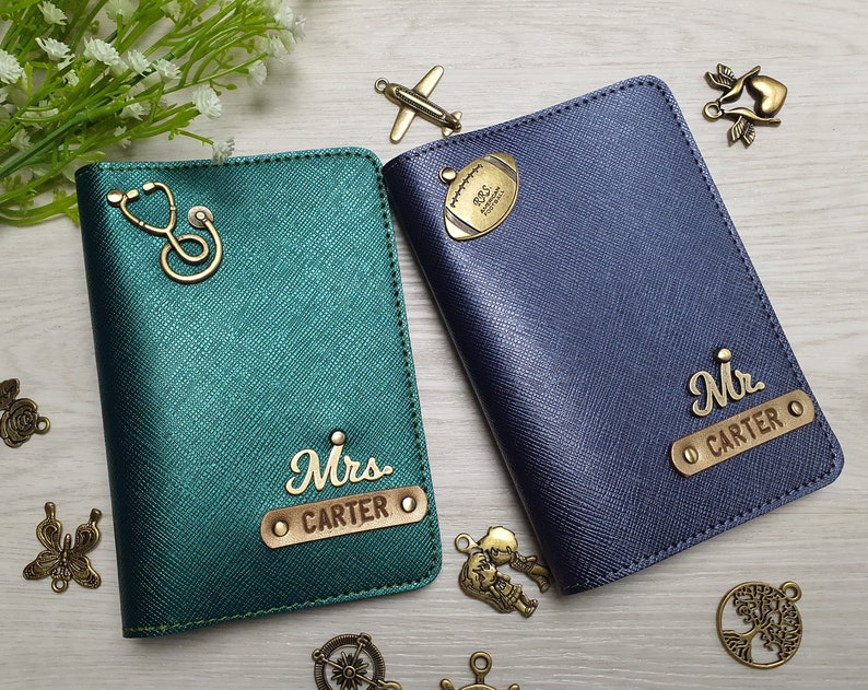 May include: Two personalized passport holders. One is teal green with a stethoscope charm and "Mrs. CARTER" in gold lettering. The other is navy blue with a football charm and "Mr. CARTER" in gold lettering. Decorative charms surround the holders.