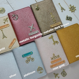 May include: A collection of passport holders in various colors, including gold, metallic burgundy, dark brown, silver, platinum, rose gold, and copper. Each holder features decorative charms and personalized nameplates. The text "WANDERLUST" and "CAMILA" are visible.