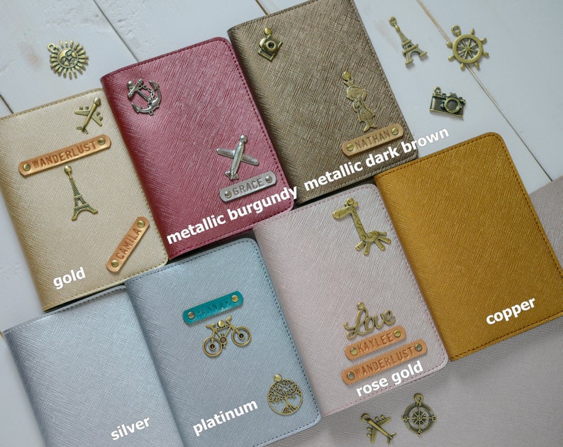 May include: A collection of passport holders in various colors, including gold, metallic burgundy, dark brown, silver, platinum, rose gold, and copper. Each holder features decorative charms and personalized nameplates. The text "WANDERLUST" and "CAMILA" are visible.