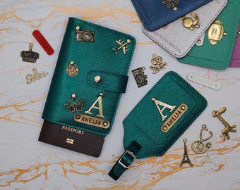 May include: Teal passport holder and luggage tag with gold accents. The passport holder has a gold letter "A" and the name "AMELIA". Decorative charms include a camera, airplane, and crown. Other luggage tags are visible.