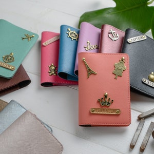 May include: A collection of colorful passport holders with decorative charms and personalized nameplates. The holders are in various colors, including teal, pink, and black. Charms include an airplane, a ship's wheel, and a crown. The nameplates have names like "William" and "Jacob".