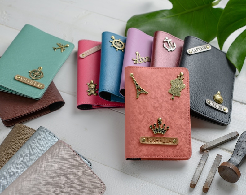 May include: A collection of colorful passport holders with decorative charms and personalized nameplates. The holders are in various colors, including teal, pink, and black. Charms include an airplane, a ship's wheel, and a crown. The nameplates have names like "William" and "Jacob".