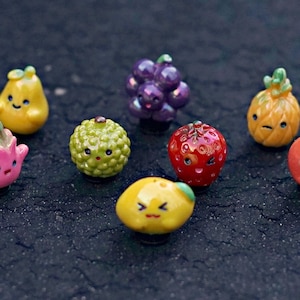 May include: A collection of colorful, miniature fruit-shaped figurines with smiling faces. The assortment includes a yellow pear, purple grapes, a red strawberry, a green kiwi, a yellow lemon, a pink flower, an orange, and a small pineapple.
