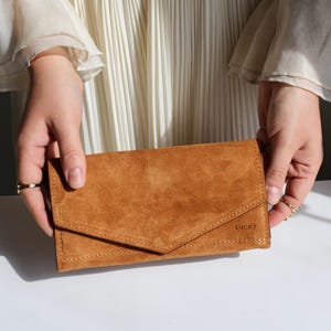 May include: A tan suede envelope-style wallet featuring a pointed flap closure and visible stitching along the edges. The word LUCKY is embossed in the bottom right corner of the front flap. The wallet has a soft, matte texture and rectangular shape.