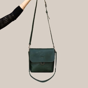 Puede incluir: Bolso bandolera de cuero verde oscuro con cierre de solapa y correa ajustable. El bolso tiene forma rectangular con un detalle de hebilla en la parte delantera. La correa mide unos 122 cm.
