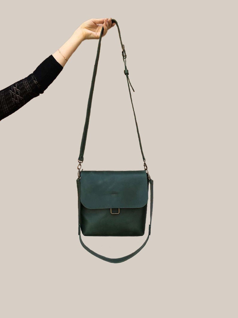 Puede incluir: Bolso bandolera de cuero verde oscuro con cierre de solapa y correa ajustable. El bolso tiene forma rectangular con un detalle de hebilla en la parte delantera. La correa mide unos 122 cm.