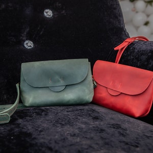 May include: Two leather crossbody bags, one green and one red, are displayed on a black velvet surface. Both bags have a flap closure and a single strap.