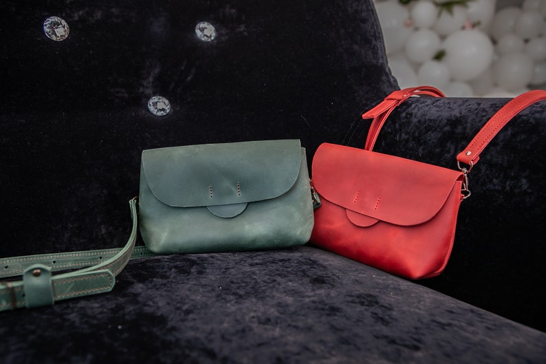 May include: Two leather crossbody bags, one green and one red, are displayed on a black velvet surface. Both bags have a flap closure and a single strap.