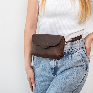 May include: A dark brown leather fanny pack with a flap closure and a small strap. The bag is worn around the waist and has a metal clasp. The leather has a distressed look.