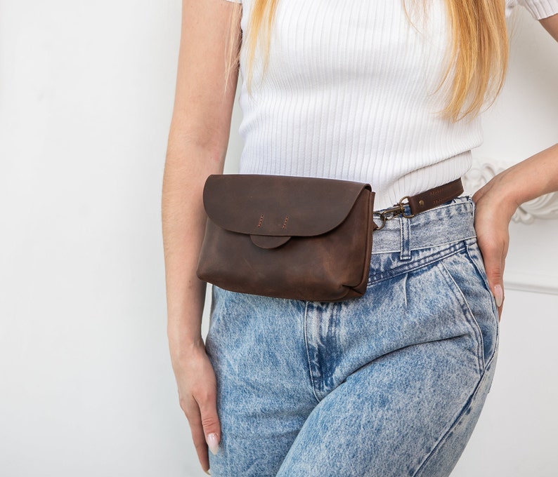 May include: A dark brown leather fanny pack with a flap closure and a small strap. The bag is worn around the waist and has a metal clasp. The leather has a distressed look.