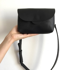 May include: A black leather crossbody bag with a curved flap closure. The bag has a long, adjustable strap and a small, circular detail on the front. The bag is held up by a hand against a white background.
