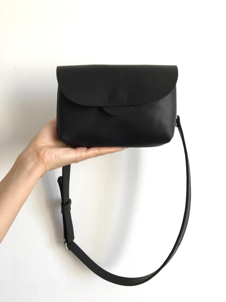 May include: A black leather crossbody bag with a curved flap closure. The bag has a long, adjustable strap and a small, circular detail on the front. The bag is held up by a hand against a white background.