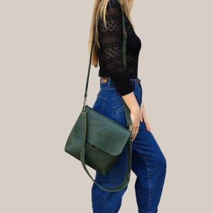 Puede incluir: Un bolso bandolera de cuero verde oscuro con cierre de solapa y correa ajustable. El bolso se lleva al hombro. El bolso se combina con unos vaqueros azules y una camisa negra de manga larga.