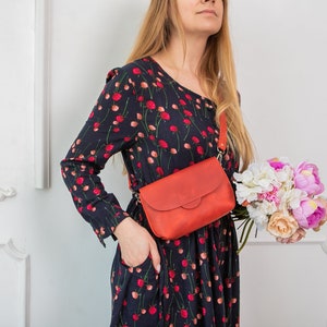 May include: A red leather crossbody bag with a flap closure is worn across the body. The bag has a long strap and is paired with a dark blue dress with a red floral pattern. A bouquet of flowers is held in the hand.