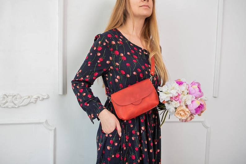 May include: A red leather crossbody bag with a flap closure is worn across the body. The bag has a long strap and is paired with a dark blue dress with a red floral pattern. A bouquet of flowers is held in the hand.