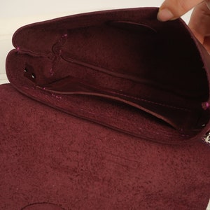 May include: Open view of a burgundy leather crossbody bag. The bag's interior reveals multiple compartments and a flap closure. The stitching is a contrasting pink color, and the bag appears to be made of soft, textured leather.