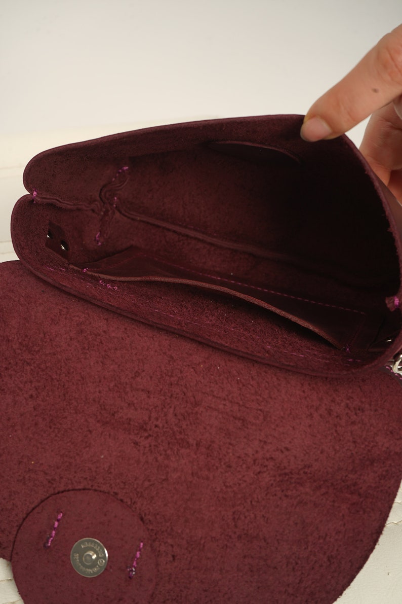 May include: Open view of a burgundy leather crossbody bag. The bag's interior reveals multiple compartments and a flap closure. The stitching is a contrasting pink color, and the bag appears to be made of soft, textured leather.
