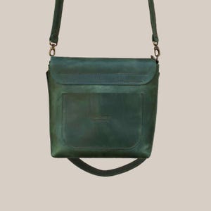 Puede incluir: Un bolso bandolera de cuero verde con cierre de solapa y bolsillo frontal. El bolso tiene una correa larga y ajustable y herrajes de color bronce. El cuero tiene un aspecto envejecido, d&aacute;ndole un aspecto vintage.