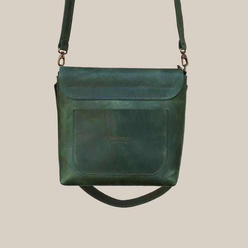 Puede incluir: Un bolso bandolera de cuero verde con cierre de solapa y bolsillo frontal. El bolso tiene una correa larga y ajustable y herrajes de color bronce. El cuero tiene un aspecto envejecido, d&aacute;ndole un aspecto vintage.