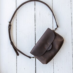 May include: A brown leather crossbody bag with a flap closure and a long adjustable strap. The bag is laying on a white wooden surface.