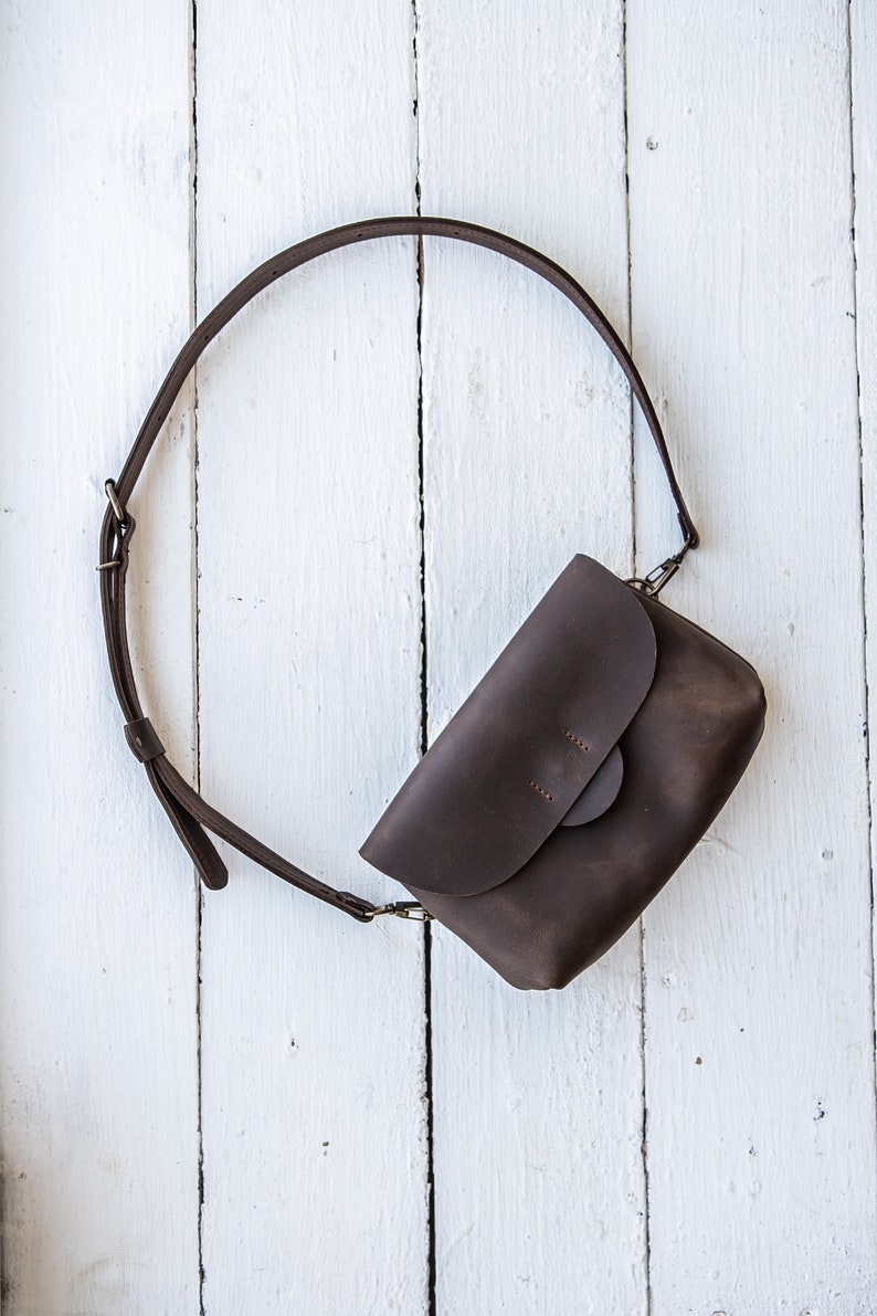 May include: A brown leather crossbody bag with a flap closure and a long adjustable strap. The bag is laying on a white wooden surface.