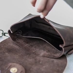 May include: A brown leather crossbody bag with a flap closure and a small pocket inside. The bag has a brown suede lining and a gold-tone buckle.