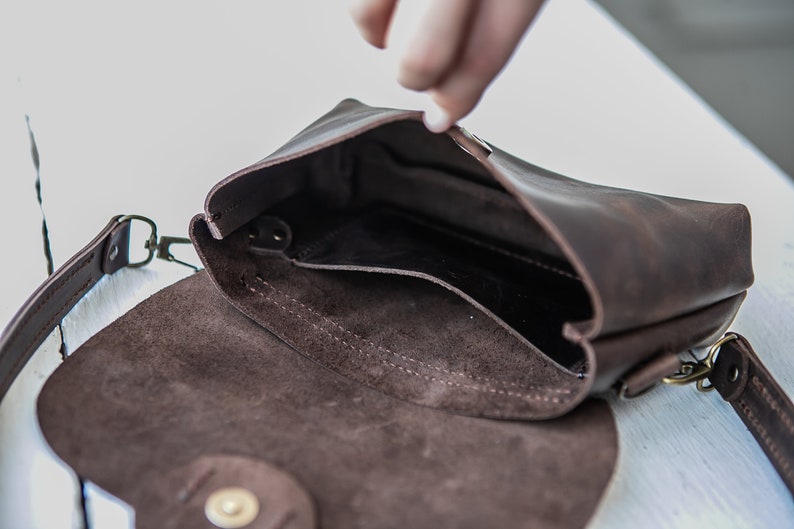 May include: A brown leather crossbody bag with a flap closure and a small pocket inside. The bag has a brown suede lining and a gold-tone buckle.