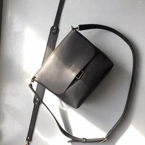 Puede incluir: Bolso bandolera de cuero gris oscuro con cierre de solapa y correa de hombro larga y ajustable. El bolso presenta herrajes dorados y una correa desmontable. El dise&ntilde;o es minimalista y moderno.